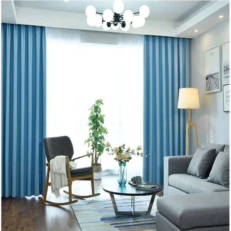 Salon contemporain avec rideaux bleus, canapé gris, et chaise berçante en bois. Table basse moderne sur tapis à motifs, lampe sur pied élégante, et décoration florale. Ambiance lumineuse et confortable.