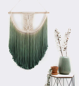 Décoration murale bohème avec dégradé de vert suspendue sur une baguette en bois, ajoutant une touche naturelle à l'intérieur. Cette tapisserie artistique complète parfaitement une ambiance zen, avec des éléments végétaux et une table d'appoint au design moderne. Idéal pour un salon ou une chambre.
