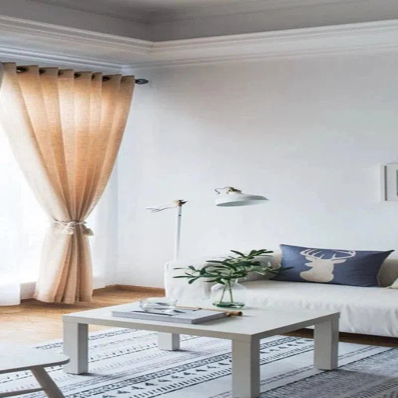 Salon moderne et lumineux avec rideaux beige, canapé élégant orné d'un coussin design motif cerf, lampe contemporaine et table basse minimaliste sur tapis à motifs géométriques. Décoration naturelle avec plante verte, créant une ambiance paisible et raffinée.