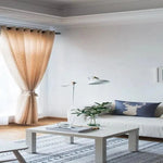 Salon moderne et lumineux avec rideaux beige, canapé élégant orné d'un coussin design motif cerf, lampe contemporaine et table basse minimaliste sur tapis à motifs géométriques. Décoration naturelle avec plante verte, créant une ambiance paisible et raffinée.