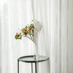 Voile décoratif élégant suspendu devant une table d'appoint verte avec un vase et une branche fleurie, ajoutant une touche de sophistication à un salon ou une chambre. Idéal pour créer une atmosphère lumineuse et naturelle tout en préservant une certaine intimité.