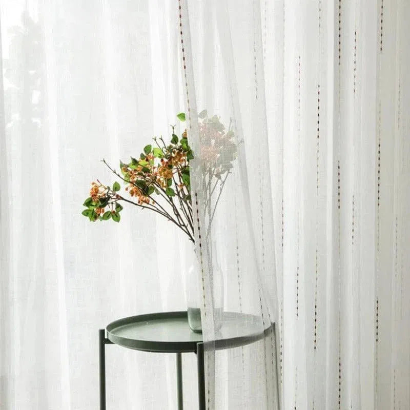Voile décoratif élégant suspendu devant une table d'appoint verte avec un vase et une branche fleurie, ajoutant une touche de sophistication à un salon ou une chambre. Idéal pour créer une atmosphère lumineuse et naturelle tout en préservant une certaine intimité.