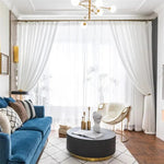 Salon contemporain et élégant avec canapé bleu, table basse ronde noire, chaise moderne et rideaux blancs légers. Décoration intérieure sophistiquée dans un espace lumineux mettant en valeur des détails dorés subtils. Idéal pour une ambiance chic et accueillante.