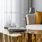 Salon moderne avec table basse dorée au design artistique, canapé crème orné de coussins colorés, et rideaux élégants en arrière-plan. Décoration intérieure chic et lumineuse, idéale pour un espace de vie accueillant et contemporain. Parfait pour ajouter une touche d’élégance à tout intérieur.