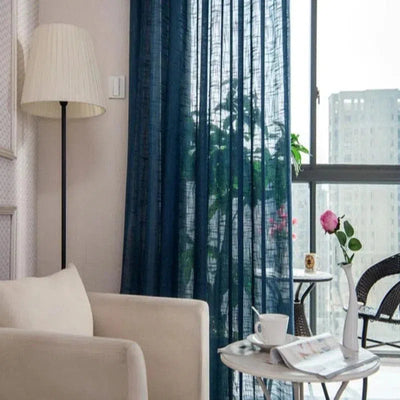 Salon lumineux et moderne avec rideaux bleus transparents soulignant une grande fenêtre. La pièce est aménagée avec un canapé confortable, une lampe élégante, et une table d'appoint décorée d'une tasse et d'un vase avec une fleur rose, créant une atmosphère accueillante et raffinée.