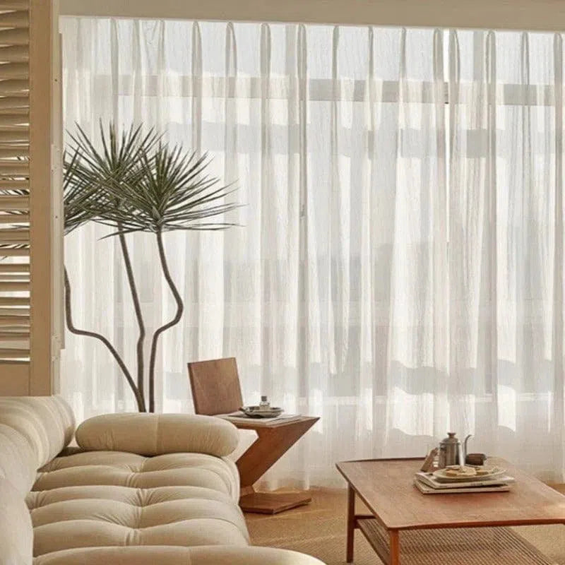 Salon moderne et lumineux avec une décoration minimaliste, offrant un espace de détente avec un canapé élégant et une table basse raffinée. Les rideaux translucides filtrent la lumière naturelle, créant une ambiance chaleureuse et apaisante. L'intérieur affiche un style scandinave épuré, propice à la relaxation.