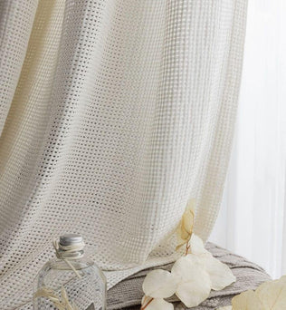Rideau opaque ivoire avec texture géométrique laissant passer la lumière. Élément de décoration idéal pour embellir un salon ou une chambre à coucher. Flacon en verre et fleurs séchées en arrière-plan pour une ambiance naturelle et apaisante.