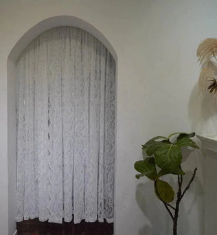 Rideau embellissant une fenêtre arquée dans un intérieur élégant avec décoration végétale. Parfait pour ajouter une touche de raffinement et de charme classique à votre pièce. Idéal pour un salon ou une chambre, apportant une lumière douce et filtrée pour une atmosphère paisible.
