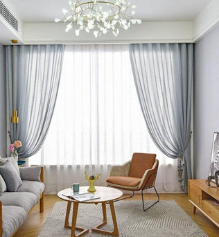 Salon moderne avec décoration élégante, comprenant un canapé confortable, un fauteuil design et une table basse en bois. Grands rideaux bleus encadrant une fenêtre lumineuse, créant une ambiance chaleureuse et accueillante. Lustre suspendu au plafond apportant une touche contemporaine à l'ensemble. Meuble de rangement en bois clair avec téléviseur moderne, idéal pour un espace de vie harmonieux et chic.