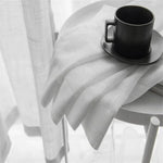 Tasse à café noire élégante sur une table blanche, posée sur une nappe immaculée, avec des rideaux lumineux en arrière-plan. Idéale pour une décoration moderne et minimaliste.