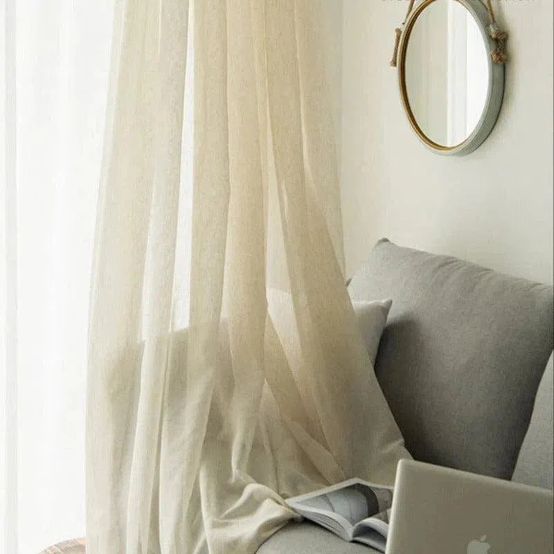Rideau beige décoratif suspendu devant une fenêtre, laissant passer la lumière naturelle dans un salon moderne et minimaliste. Canapé gris confortable avec coussins, livre ouvert et ordinateur portable, créant un espace chaleureux et accueillant. Ensemble harmonieux pour une décoration intérieure élégante et apaisante.