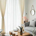 Salon moderne avec un canapé gris et coussins assortis, table basse en bois au design unique, et rideaux blancs laissant entrer la lumière naturelle. Décoration épurée avec miroir rond mural, vase avec fleurs séchées et objets déco sur meuble d'appoint. Atmosphère lumineuse et accueillante parfaite pour un intérieur contemporain.