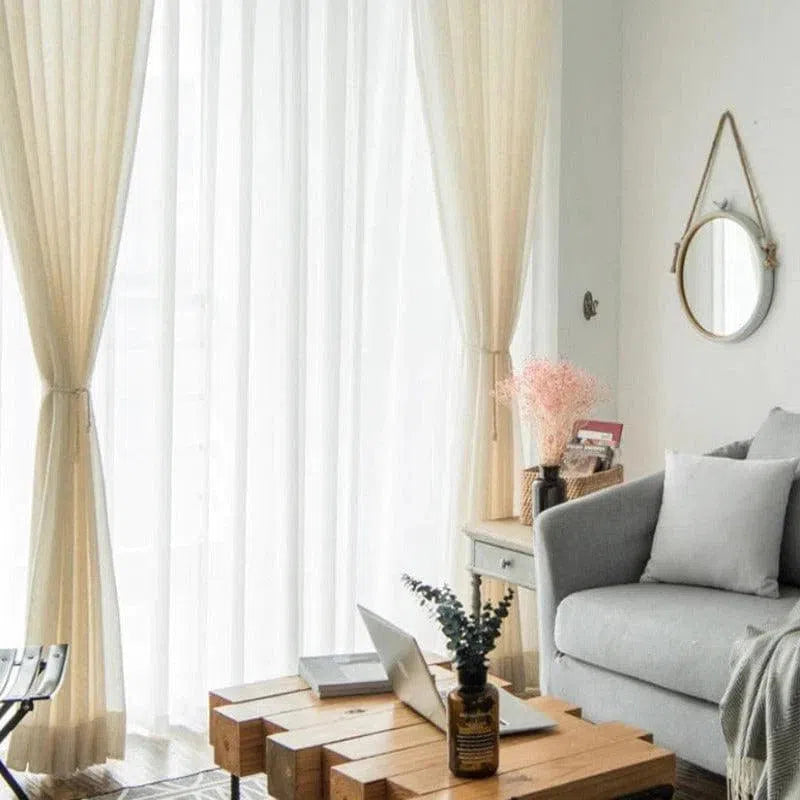 Salon moderne avec un canapé gris et coussins assortis, table basse en bois au design unique, et rideaux blancs laissant entrer la lumière naturelle. Décoration épurée avec miroir rond mural, vase avec fleurs séchées et objets déco sur meuble d'appoint. Atmosphère lumineuse et accueillante parfaite pour un intérieur contemporain.