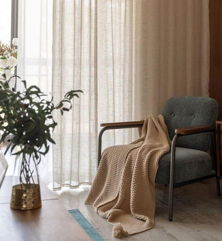 Fauteuil confortable avec plaid dans un salon lumineux, idéal pour la détente. Rideaux élégants laissant passer la lumière naturelle, plante d'intérieur et décoration moderne pour un espace de vie accueillant et chaleureux.