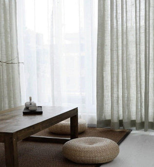 Rideaux élégants pour salon moderne, apportant une touche de luminosité et de douceur naturelle. Ambiance zen avec table basse en bois et coussins de sol tressés, créant un espace chaleureux et accueillant. Parfait pour une décoration intérieure raffinée et minimaliste.