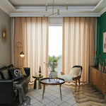 Salon moderne et élégant avec canapé en cuir noir, table basse ronde en bois et chaises d'appoint. Rideaux beiges devant une grande fenêtre apportant une lumière naturelle abondante. Décoration intérieure sophistiquée avec des plantes vertes et des lampes de style contemporain, parfait pour une ambiance chaleureuse et accueillante.