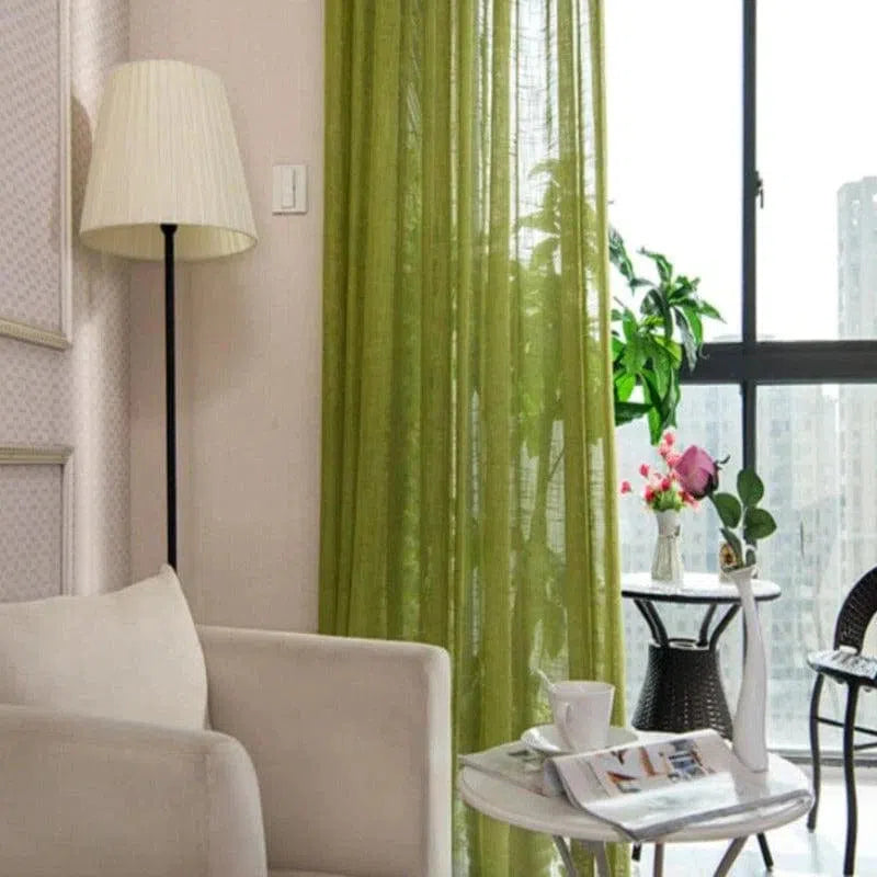 Salon moderne et lumineux avec rideaux verts, éclairage naturel grâce à une grande fenêtre. Décoration élégante avec une lampe sur pied, un canapé confortable, et une table d'appoint agrémentée de plantes et d'un vase de fleurs. Ambiance cosy et accueillante idéale pour la détente.