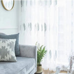 Salon moderne avec une ambiance apaisante, mettant en vedette un canapé confortable de couleur gris clair agrémenté de coussins décoratifs. Les rideaux légers avec un motif de feuilles ajoutent une touche naturelle et élégante à la pièce, filtrant doucement la lumière du jour. Un miroir rond et des plantes vertes complètent harmonieusement la décoration intérieure, créant un espace accueillant et lumineux idéal pour se détendre ou recevoir des invités.