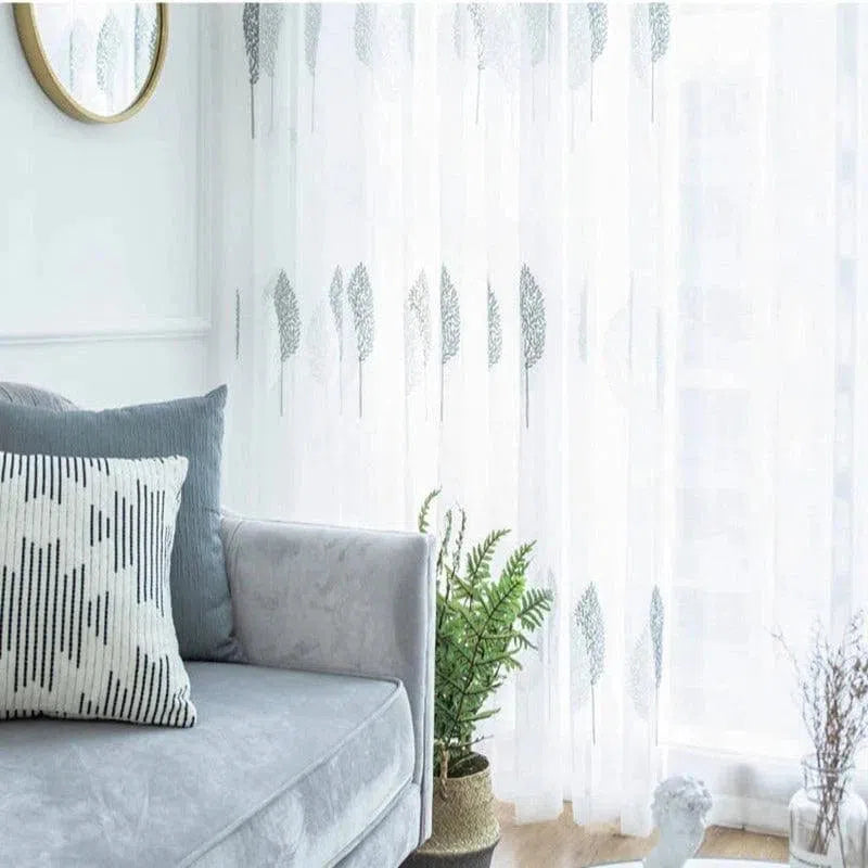 Salon moderne avec une ambiance apaisante, mettant en vedette un canapé confortable de couleur gris clair agrémenté de coussins décoratifs. Les rideaux légers avec un motif de feuilles ajoutent une touche naturelle et élégante à la pièce, filtrant doucement la lumière du jour. Un miroir rond et des plantes vertes complètent harmonieusement la décoration intérieure, créant un espace accueillant et lumineux idéal pour se détendre ou recevoir des invités.