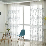 Rideaux translucides à motif floral suspendus devant une grande baie vitrée, laissant passer la lumière tout en offrant une intimité élégante. Parfait pour ajouter une touche moderne et naturelle à votre salon ou espace de travail. La chaise bleue et la table ronde complètent harmonieusement le décor lumineux et épuré.