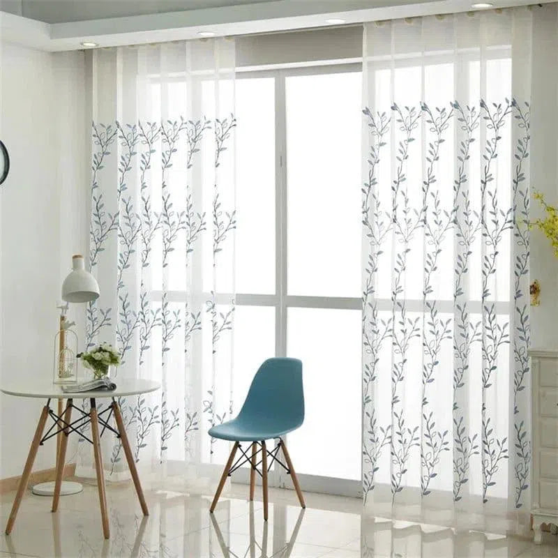 Rideaux translucides à motif floral suspendus devant une grande baie vitrée, laissant passer la lumière tout en offrant une intimité élégante. Parfait pour ajouter une touche moderne et naturelle à votre salon ou espace de travail. La chaise bleue et la table ronde complètent harmonieusement le décor lumineux et épuré.