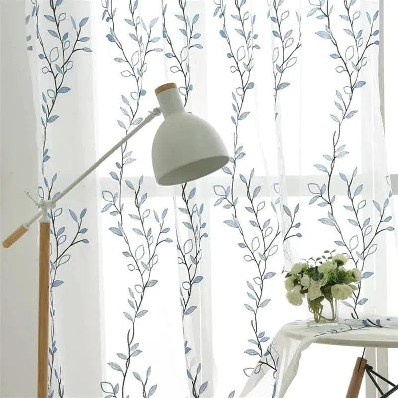 Rideaux décoratifs avec motifs floraux bleu et blanc, parfaits pour illuminer une pièce et ajouter une touche de style moderne. Accessoire idéal pour salons et chambres à coucher, qui apporte une ambiance fraîche et élégante. Lampadaire moderne en arrière-plan pour un éclairage doux et harmonieux. Vases avec fleurs blanches pour un décor raffiné et naturel.