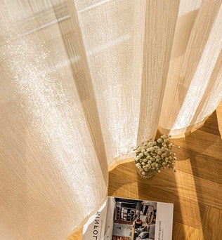Rideau lumineux offrant une ambiance apaisante avec lumière naturelle filtrée, posé sur un sol en bois chaleureux. Décoration intérieure sophistiquée avec une touche de nature, grâce à un bouquet de fleurs blanches délicatement disposé. Livre ouvert sur des idées de design moderne.