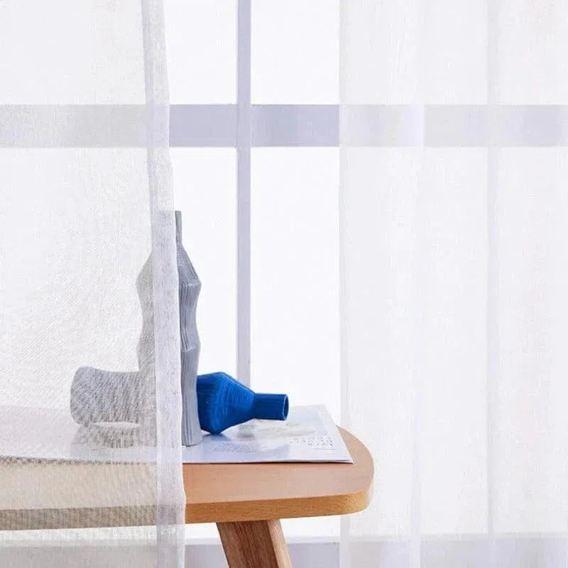 Voilage élégant et contemporain près d'une fenêtre ensoleillée, offrant un éclairage naturel et subtil. Le mobilier en bois clair ajoute de la chaleur à l'espace, tandis que des objets décoratifs apportent une touche de couleur moderne. Solution idéale pour créer une ambiance lumineuse et accueillante dans votre intérieur.