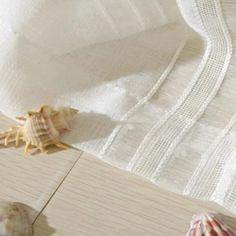 Drap blanc léger disposé sur un sol en bois clair, décoré avec des coquillages marins. Ambiance estivale et bord de mer, parfait pour une décoration intérieure fraîche et naturelle. Accessoire idéal pour apporter une touche de douceur et d'élégance à votre espace.