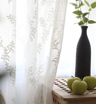 Rideau translucide élégant avec motif floral, laissant passer une douce lumière naturelle. Décoration moderne avec vase noir et pommes vertes sur une table en bois, créant une ambiance apaisante et raffinée pour la maison.